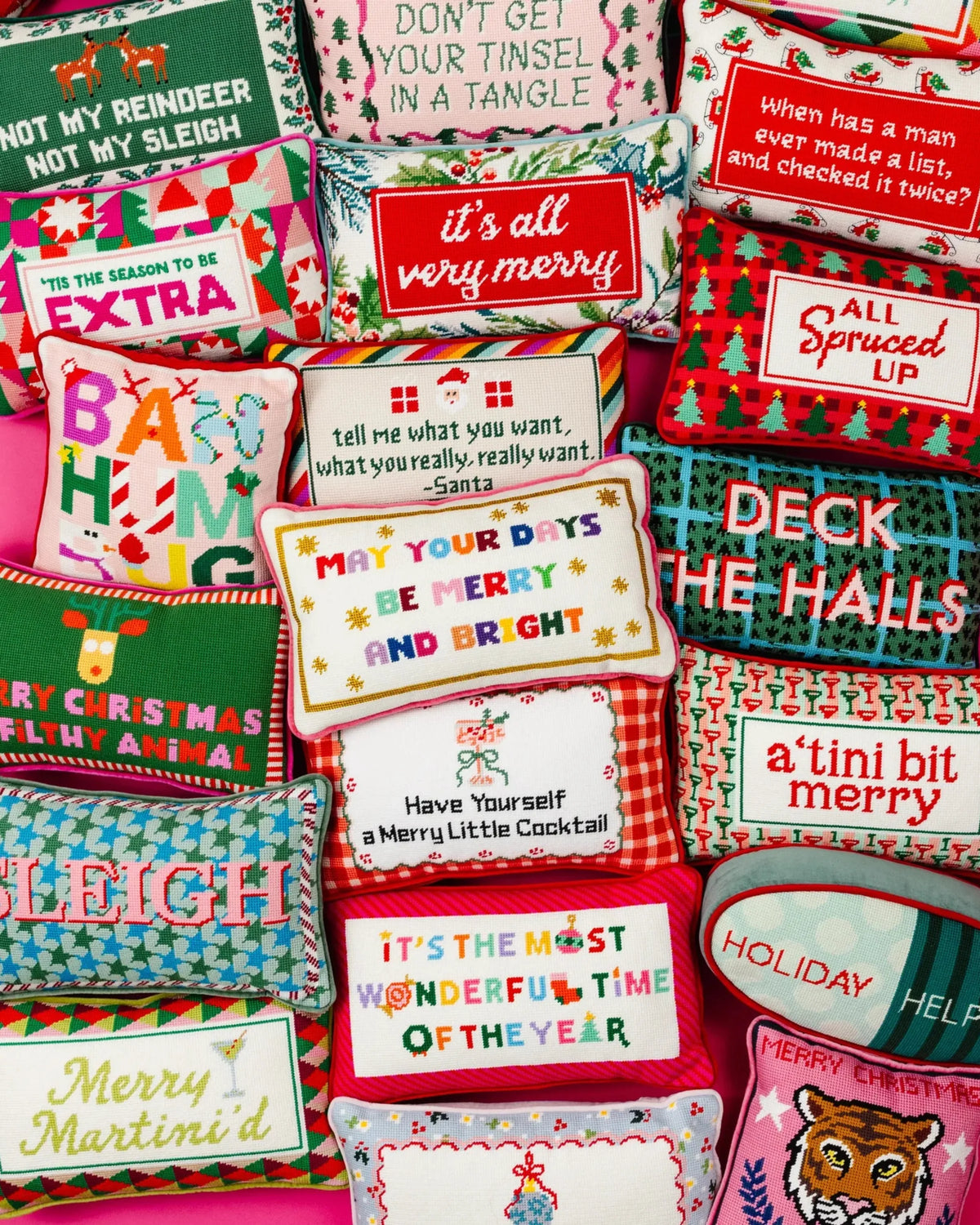 A collection of colorful Christmas-themed decorative throw pillows arranged together on a pink background, each with unique embroidered patterns and holiday quotes.