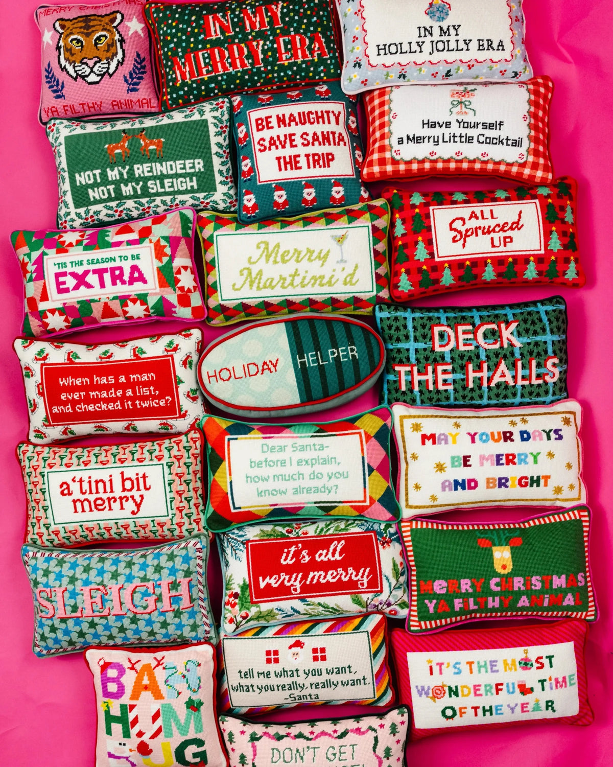 A flat-lay arrangement of multiple colorful Christmas-themed decorative pillows on a bright pink background. Each pillow features a different festive design, pattern, and humorous holiday message.