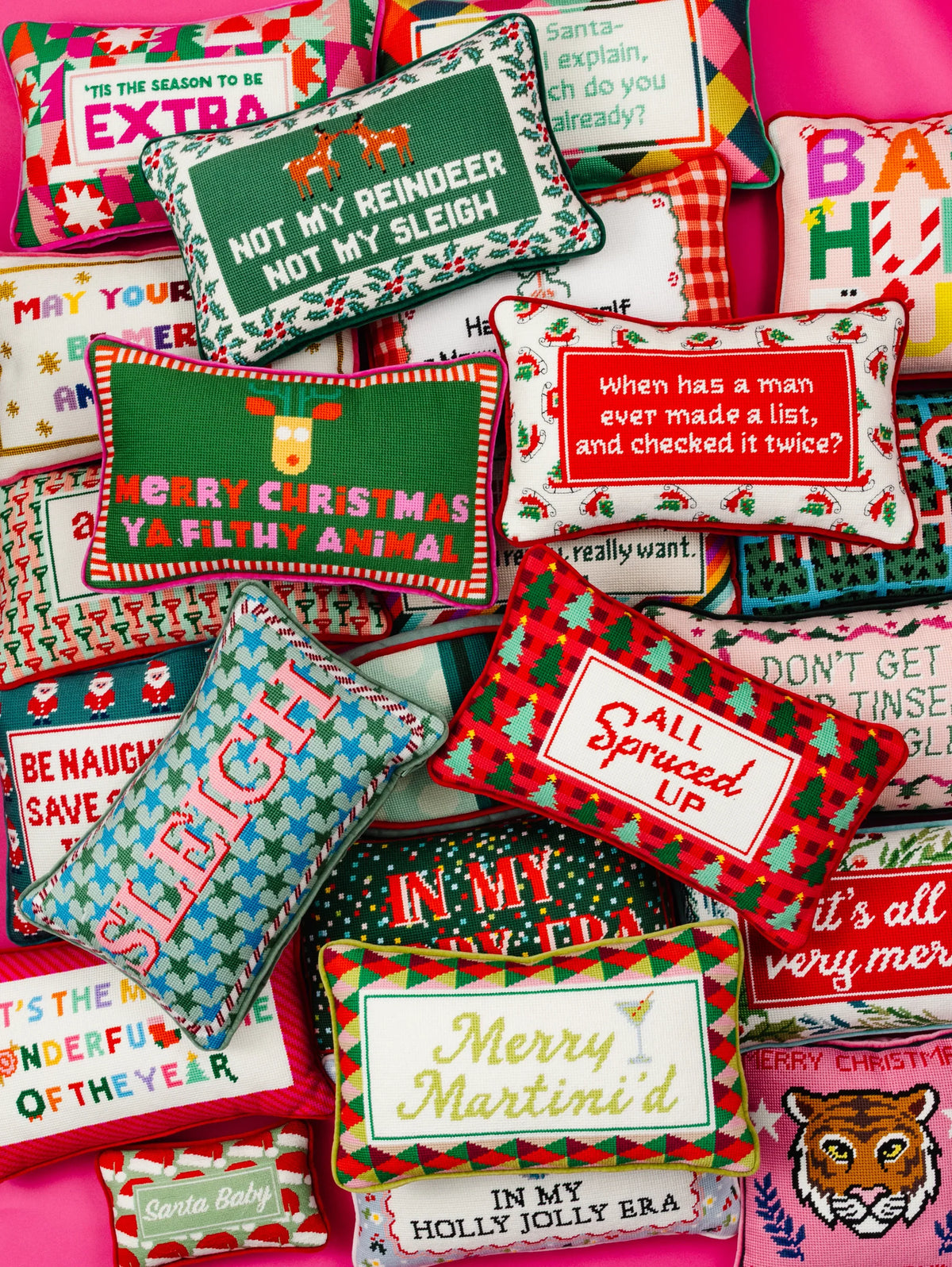 A collection of colorful Christmas-themed throw pillows with various humorous and festive printed designs, arranged on a bright pink background.