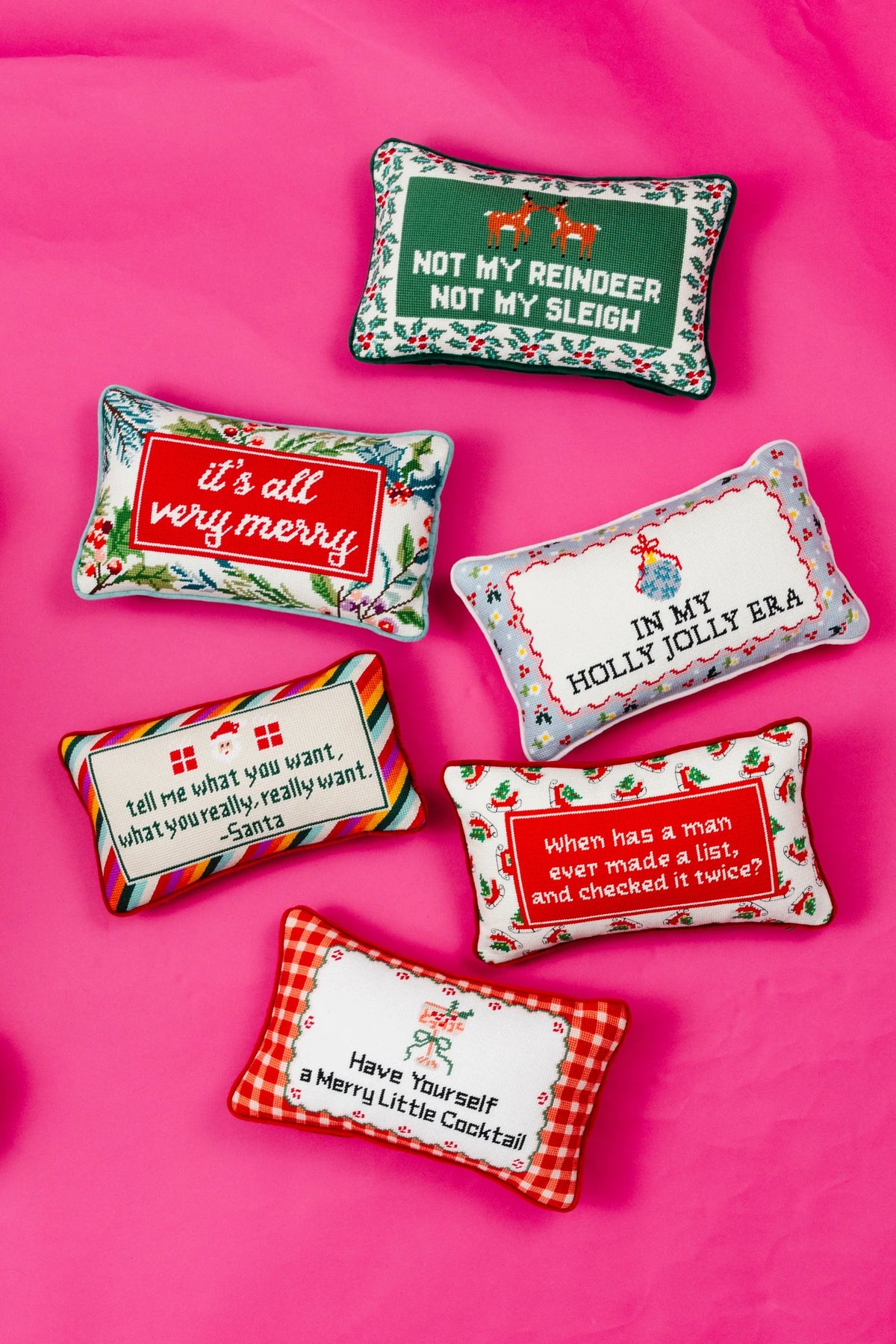 Six decorative rectangular throw pillows arranged on a bright pink fabric background. The pillows feature holiday-themed designs, including a green reindeer sign, a red 'Merry' patch, a white 'Holly Jolly Era' banner, a striped 'Santa' pillow, a red text pillow, and a red-and-white gingham 'Cocktail' pillow.
