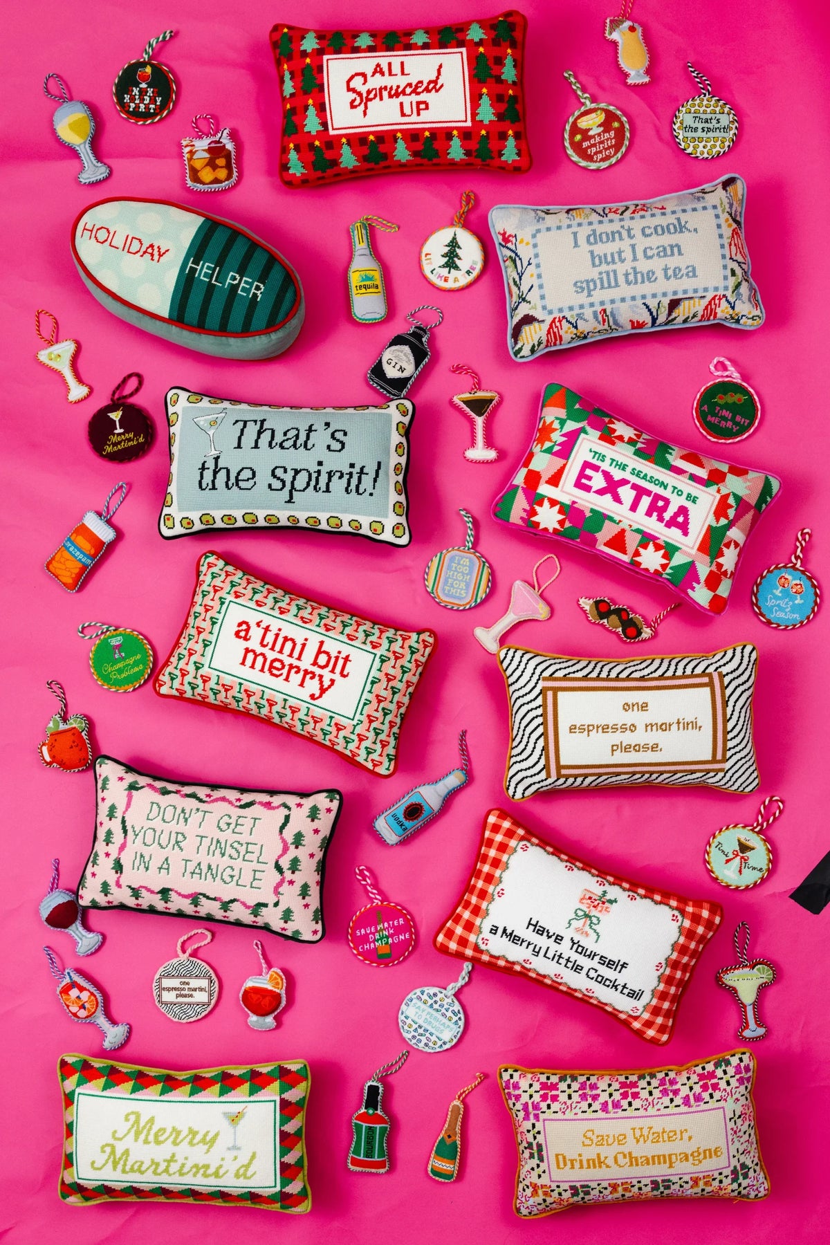 A flat-lay arrangement of multiple embroidered Christmas-themed pillows on a bright pink background, surrounded by small matching embroidered ornaments and accessories.