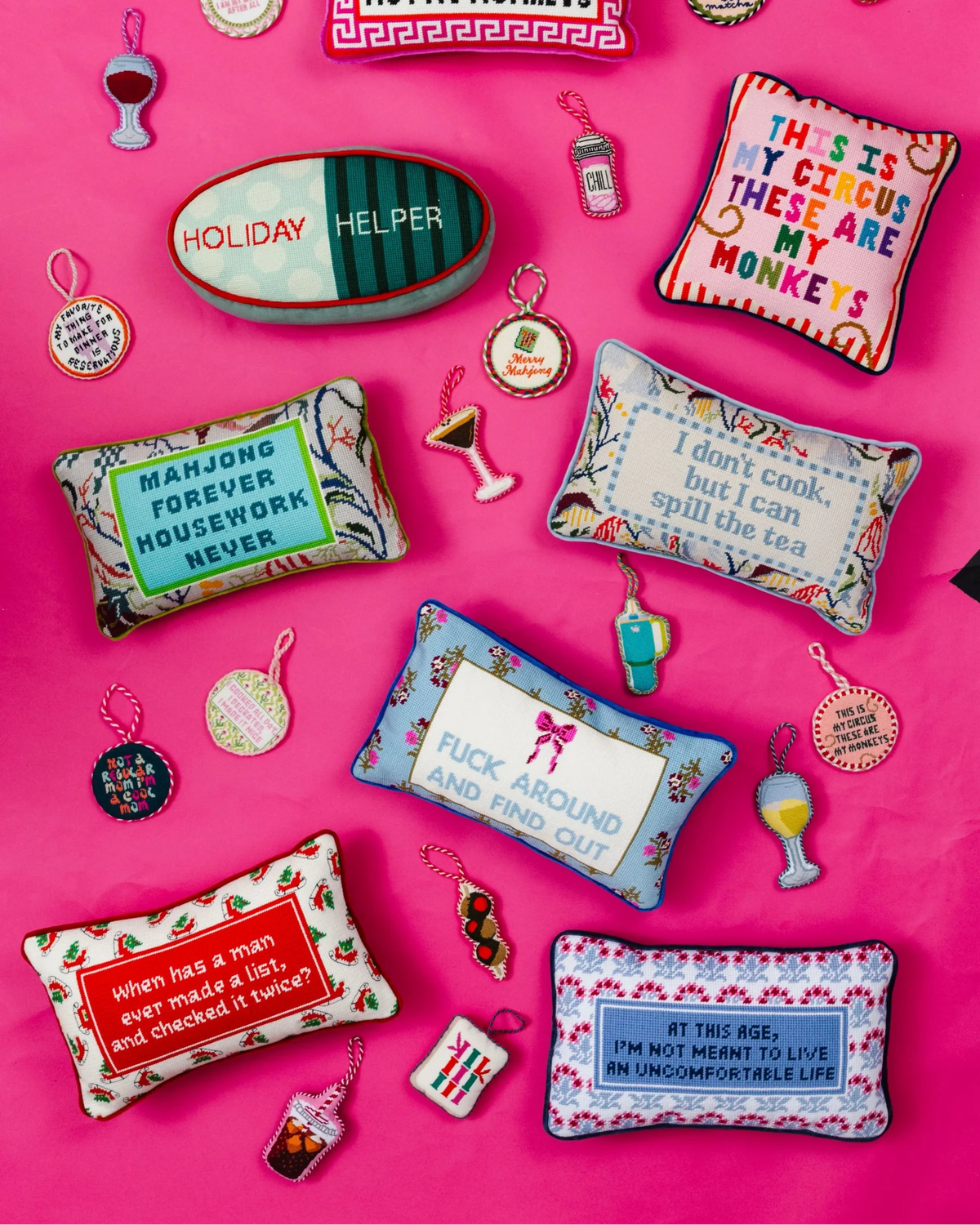 A flat lay of colorful embroidered throw pillows and small matching accessories arranged on a bright pink background. The pillows feature humorous text quotes, floral and geometric patterns, and small embroidered details like cocktail glasses, bottles, and fruit illustrations.