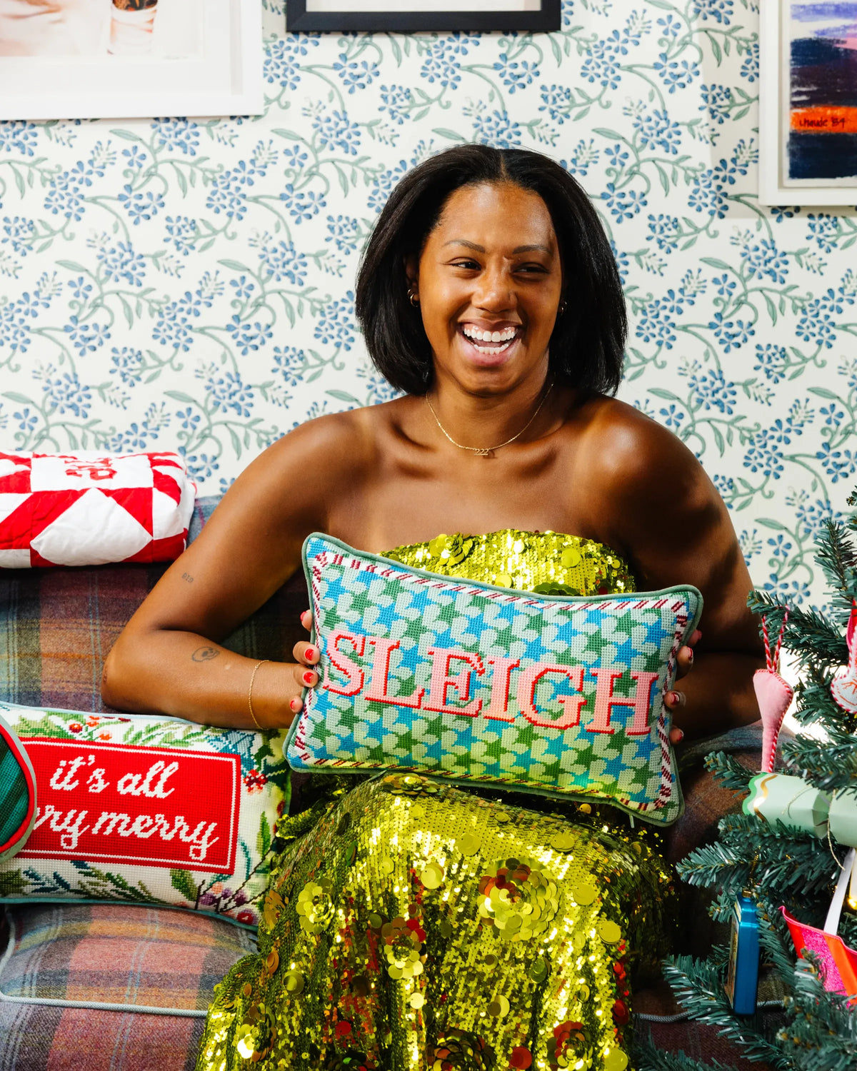 A woman smiling while holding a green and blue patterned pillow with 'SLYE' embroidered in red. She is wearing a shiny gold sequin dress, seated on a plaid couch against a blue floral wallpaper background, with other holiday pillows and a Christmas tree visible.