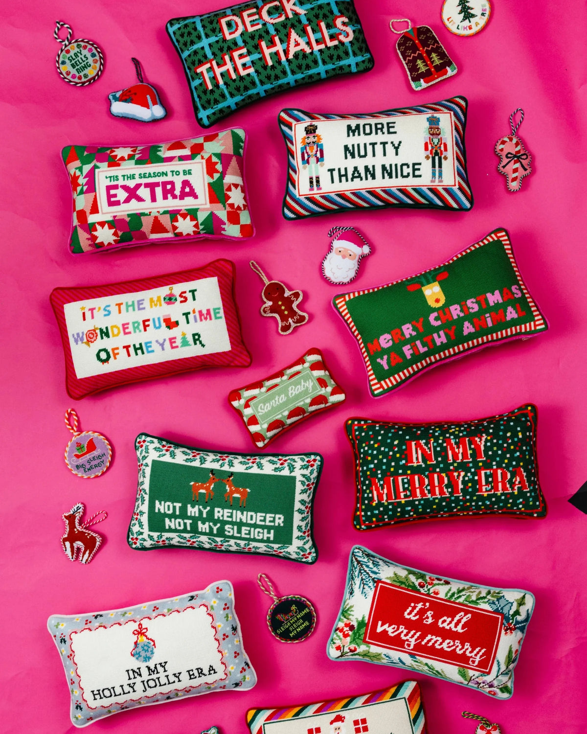 A flat lay of multiple Christmas-themed decorative throw pillows and small ornaments arranged on a bright pink background. The pillows feature festive patterns and humorous holiday sayings in red, green, white, black, and other colors.