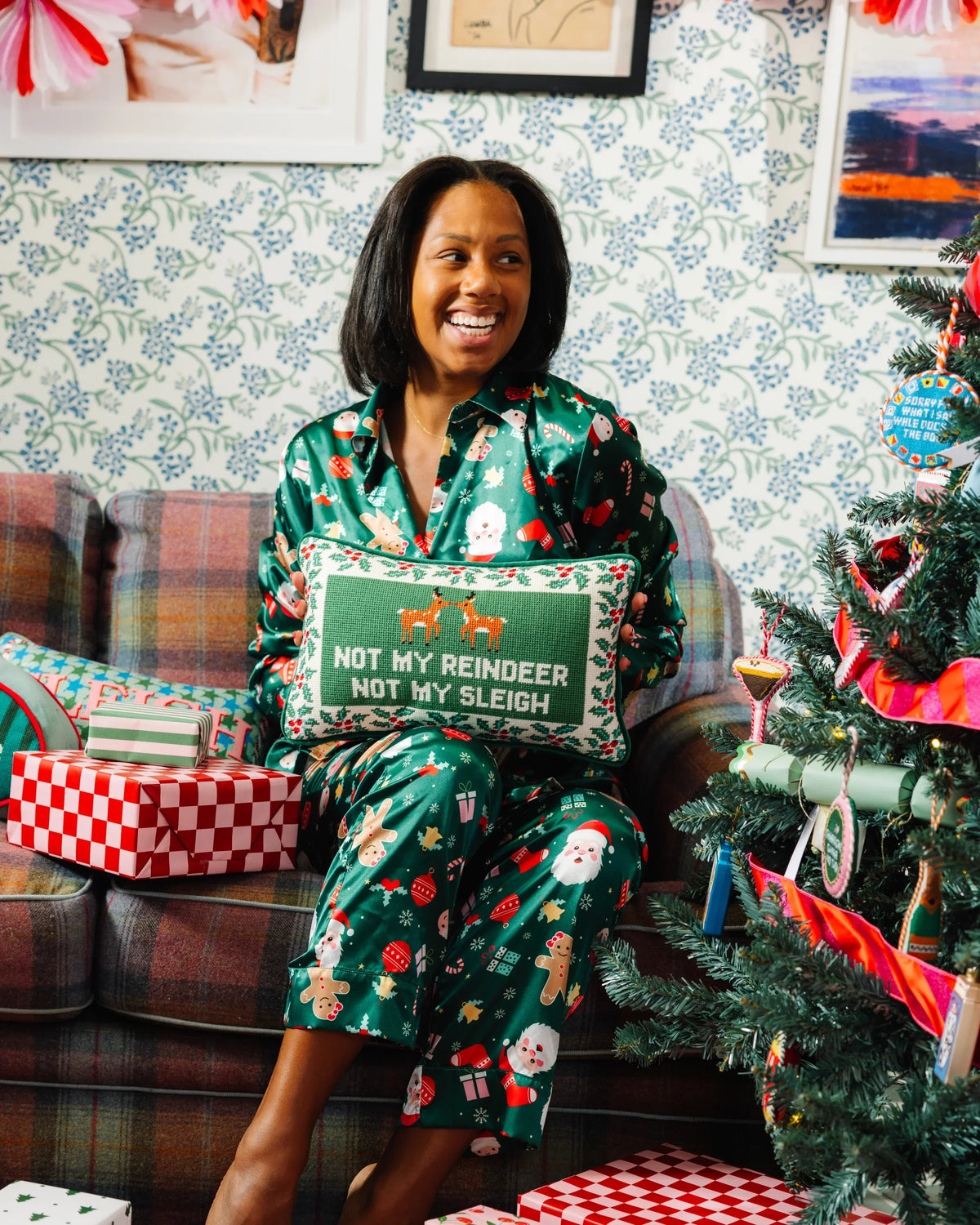 A woman wearing green Christmas-themed pajamas sits on a plaid couch next to a decorated Christmas tree, holding a decorative pillow that reads 'NOT MY REINDEER NOT MY SLEIGH'.