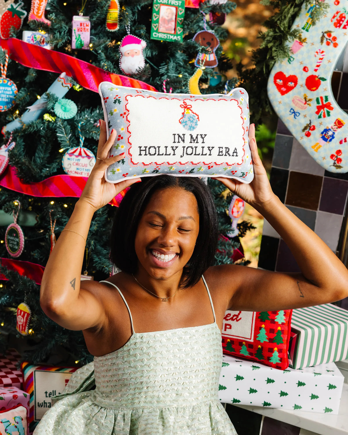 A woman in a light green patterned dress holds up a square decorative pillow with the text 'IN MY HOLLY JOLLY ERA' in front of a decorated Christmas tree and stockings.