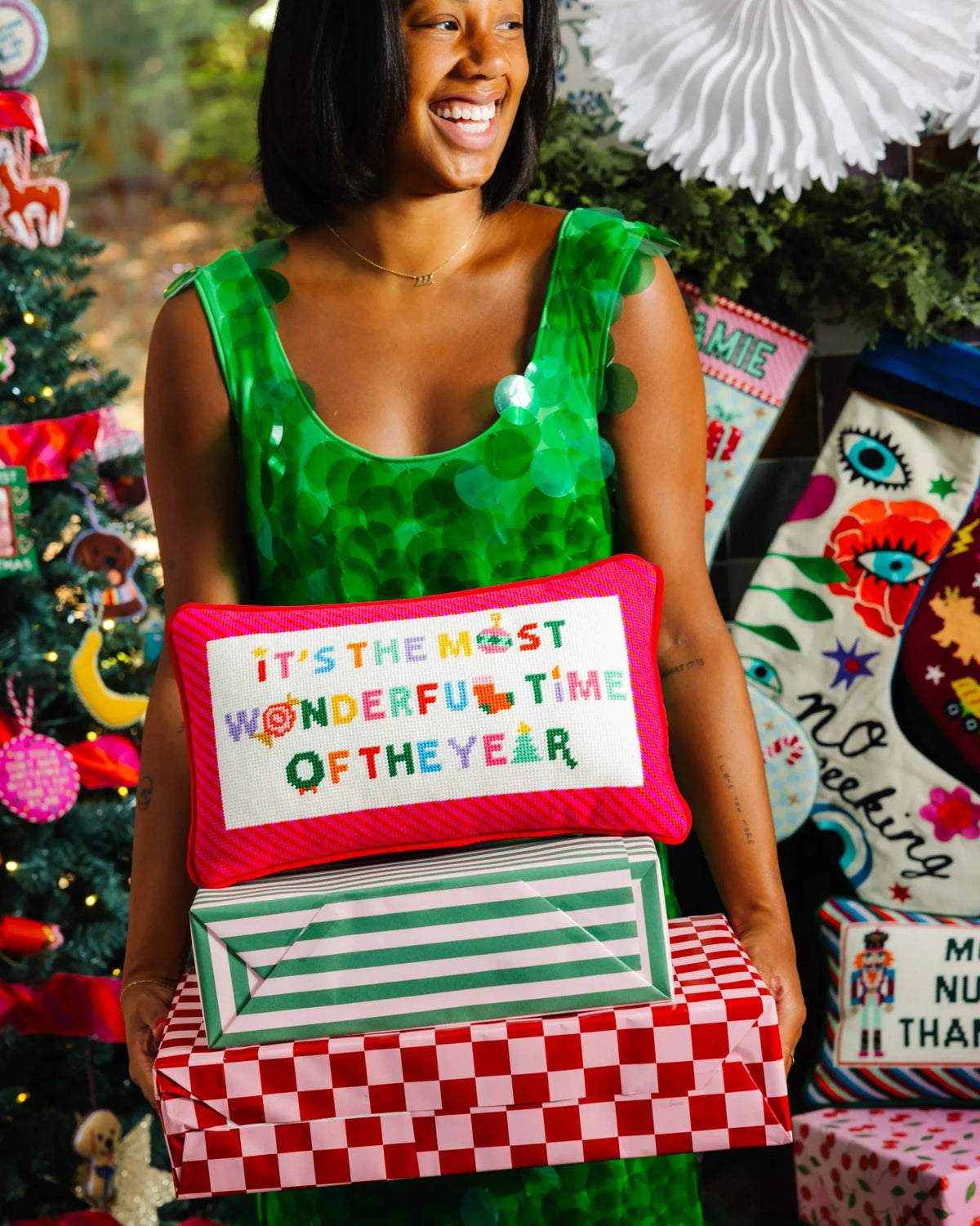 A woman in a shiny green sequined dress holds a stack of Christmas-themed gift boxes and pillows. The front pillow reads 'IT'S THE MOST WONDERFUL TIME OF THE YEAR' in colorful letters on a white background with a pink border.