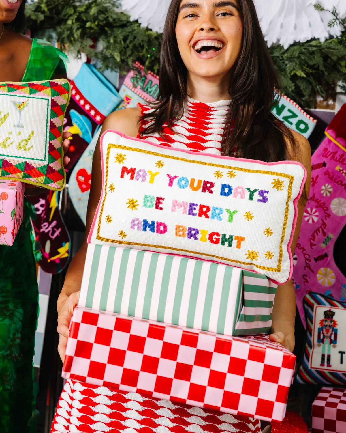 A woman smiling while holding a stack of colorful Christmas gift boxes and pillows. The central pillow reads 'MAY YOUR DAYS BE MERRY AND BRIGHT' in multicolored letters with yellow stars.