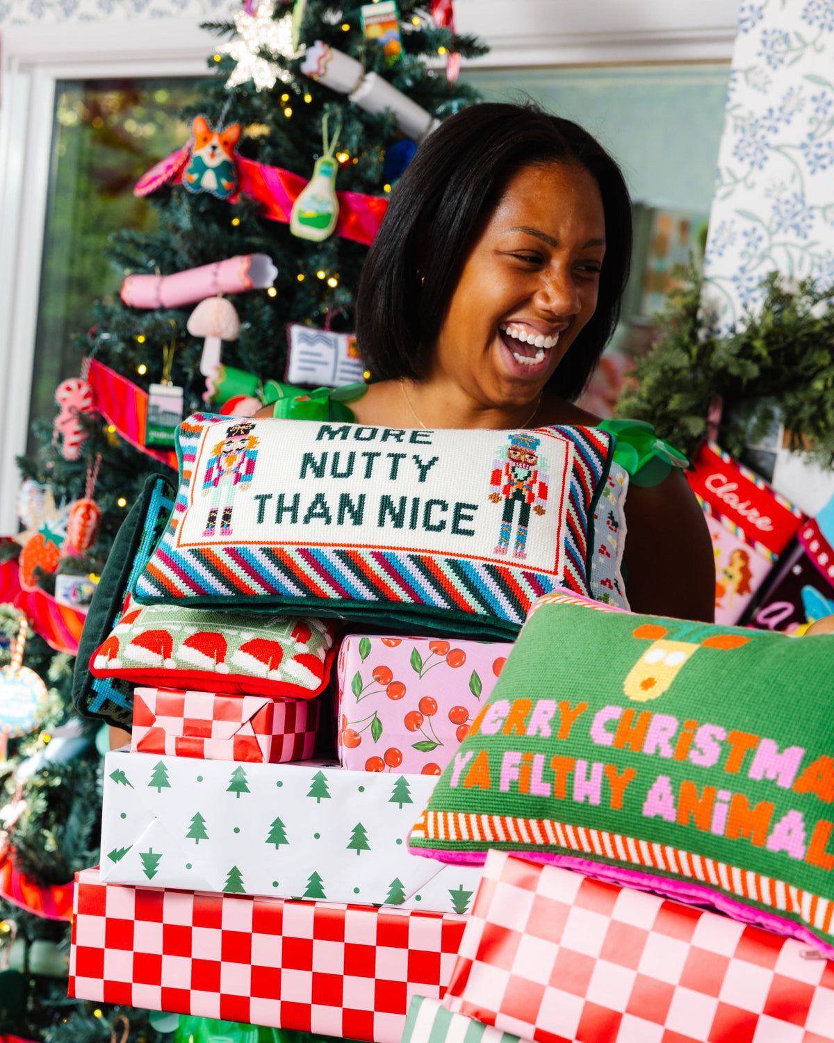 A woman laughing behind a stack of colorful Christmas-themed throw pillows and wrapped gift boxes in front of a decorated Christmas tree.