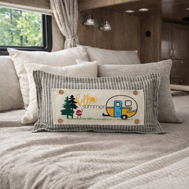 A rectangular decorative pillow with a gray and white striped border and a cream center panel, placed on a bed in a camper or RV interior. The center panel shows the words “hello summer” with a small camping trailer, tree, sun, and mushroom illustration.