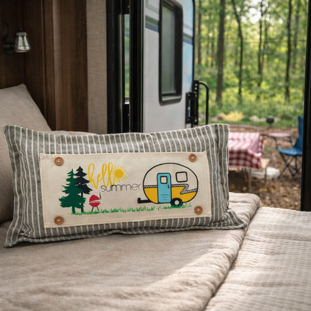 A rectangular throw pillow with a gray and white striped border and a central cream patch featuring the words 'hello summer' and camping illustrations, resting on a bed in a cozy bedroom setting.