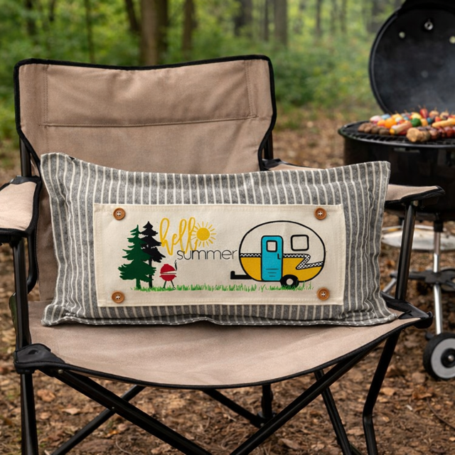 A rectangular throw pillow with a striped gray and white border and a central cream patch reading 'hello summer' with a camper, trees, and grill, resting on a beige folding camp chair outdoors.