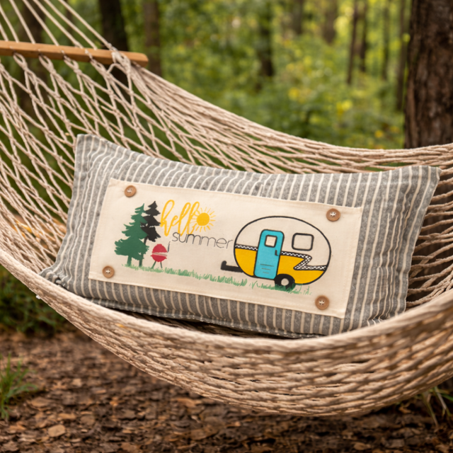 A rectangular decorative pillow with a gray and white striped fabric body and a central cream patch featuring a retro camper illustration and the words 'fall summer,' resting on a woven rope hammock in a wooded outdoor setting.