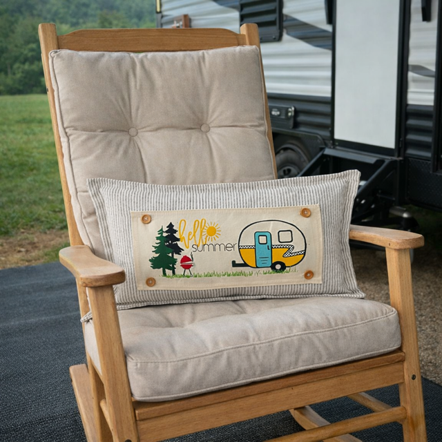 A rectangular lumbar pillow with a striped fabric border and a central graphic reading 'hello summer' with a camper trailer, trees, and a mushroom, resting on a wooden rocking chair outdoors with an RV in the background.