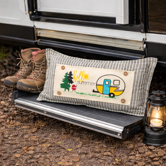 A rectangular decorative throw pillow with a striped gray and white fabric body and a central cream patch reading 'fall summer' with a camper, trees, and grill, styled on the step of an RV beside hiking boots and a lantern.