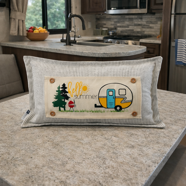 A rectangular striped throw pillow with a central embroidered patch reading 'hello summer' and showing a retro trailer, tree, and mushroom, placed on a kitchen island.
