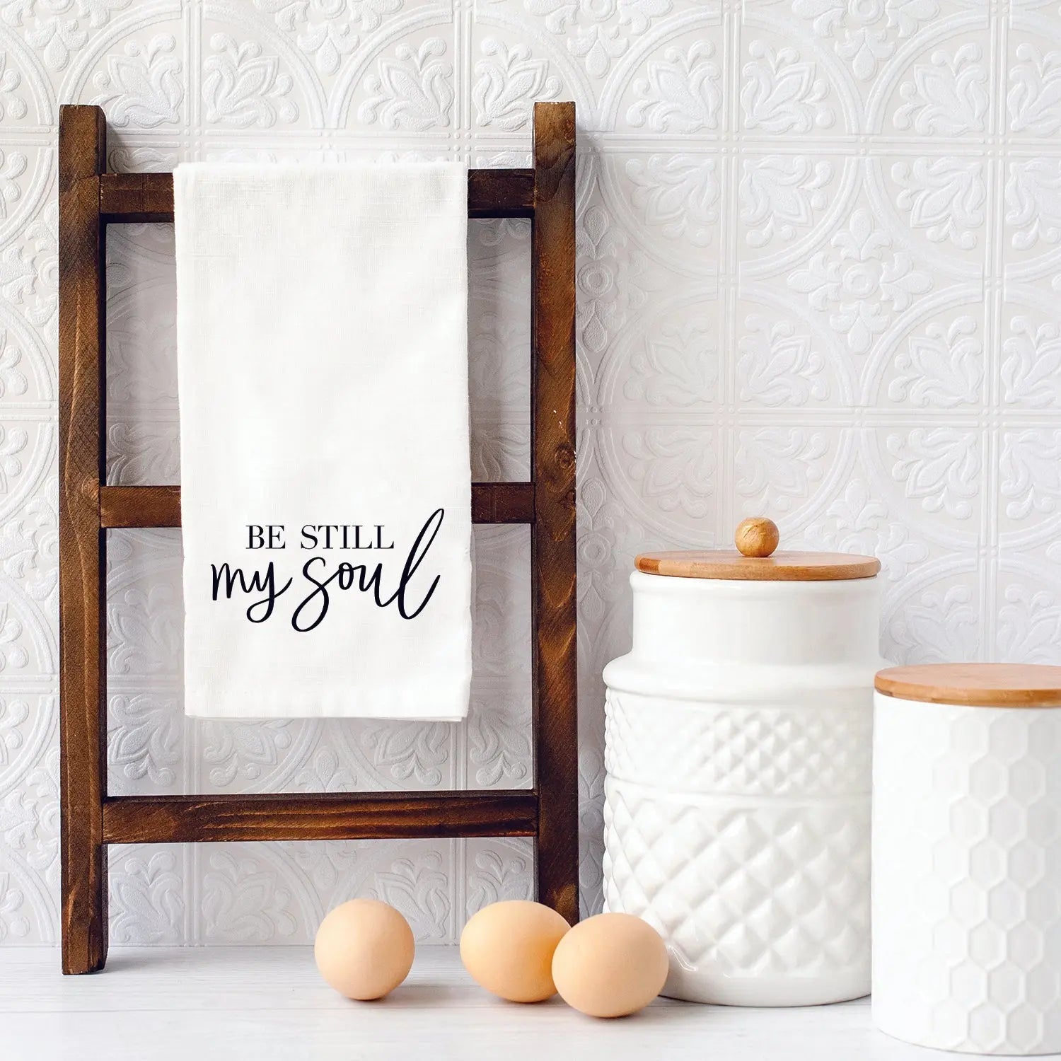 White farmhouse kitchen towel with 'Be Still My Soul' text on wooden ladder, cozy cottage decor