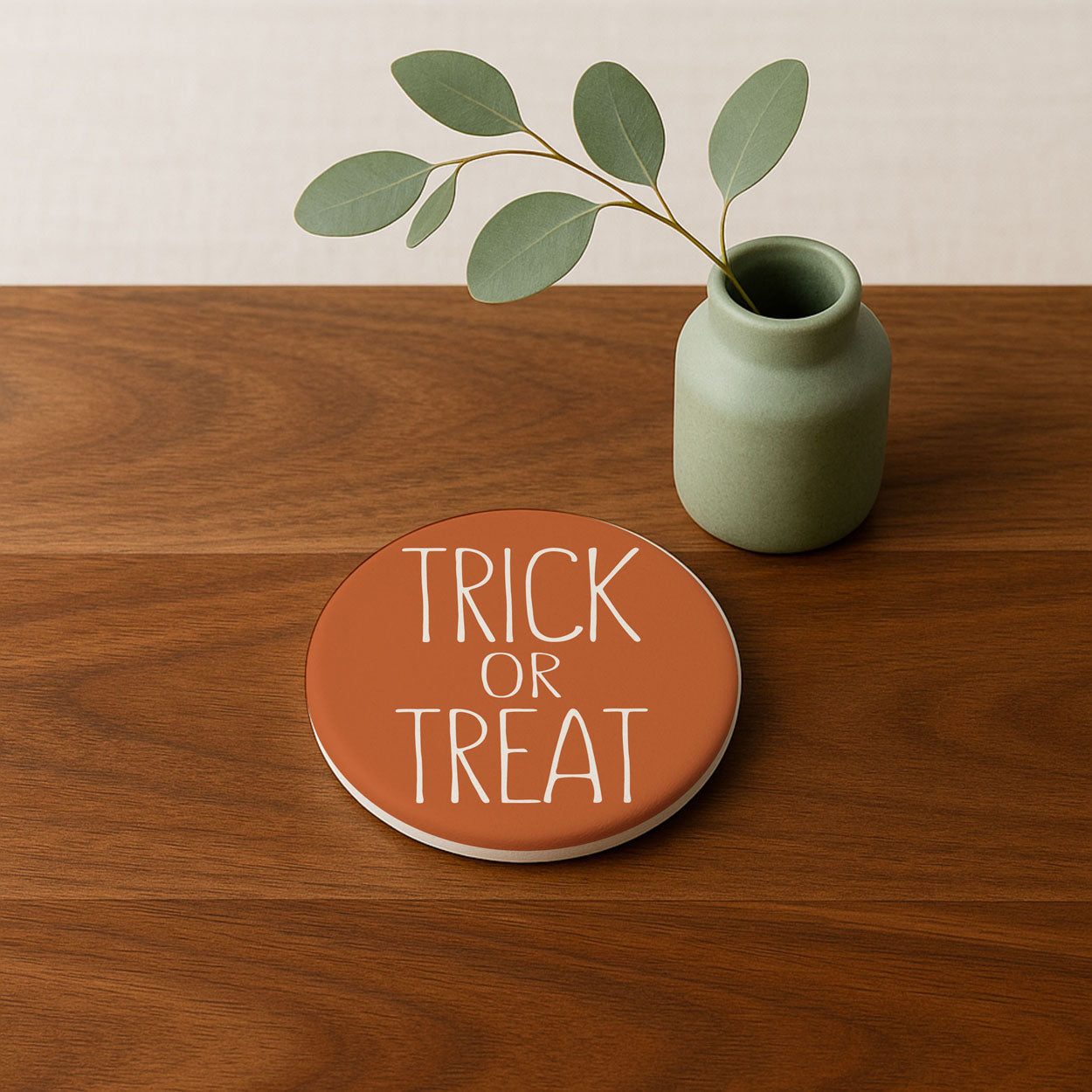 Round 'Trick or Treat' coaster on a wooden surface with a green vase and leaves.