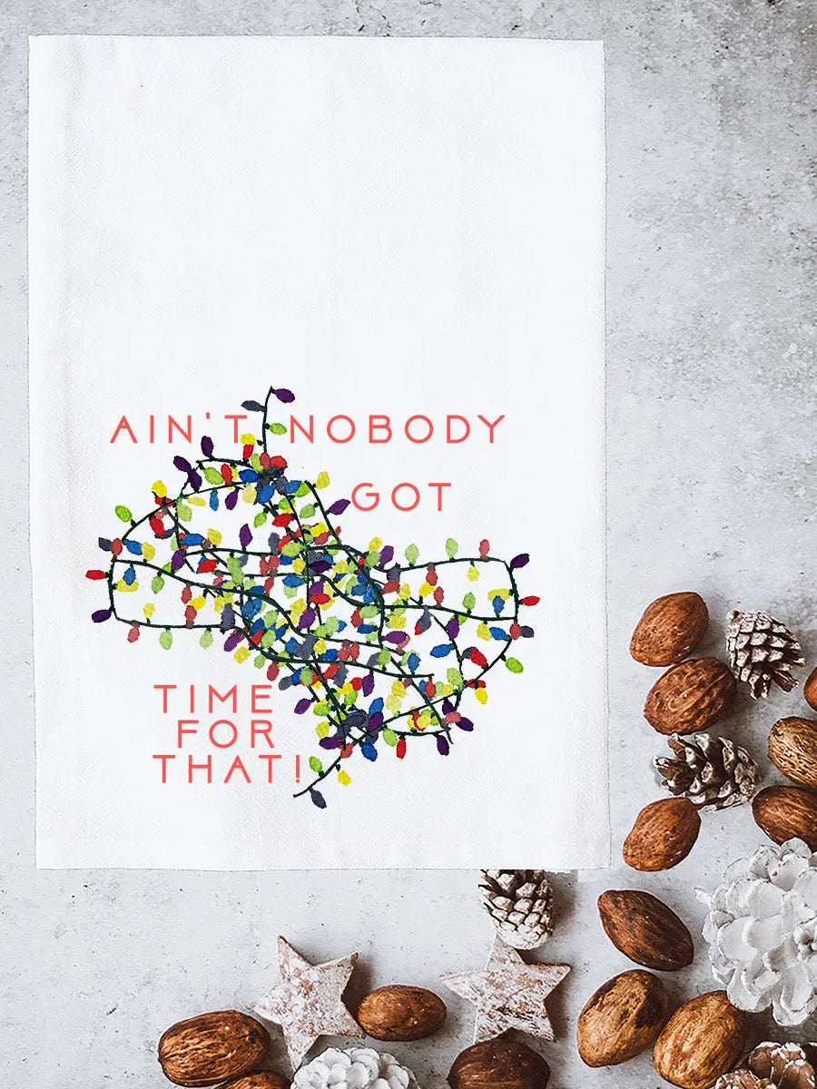 A white kitchen towel with a colorful Christmas string lights graphic and the text 'AIN'T NOBODY GOT TIME FOR THAT!' in coral pink, styled in a flat lay with pinecones, walnuts, and white decorative stars on a gray concrete surface.