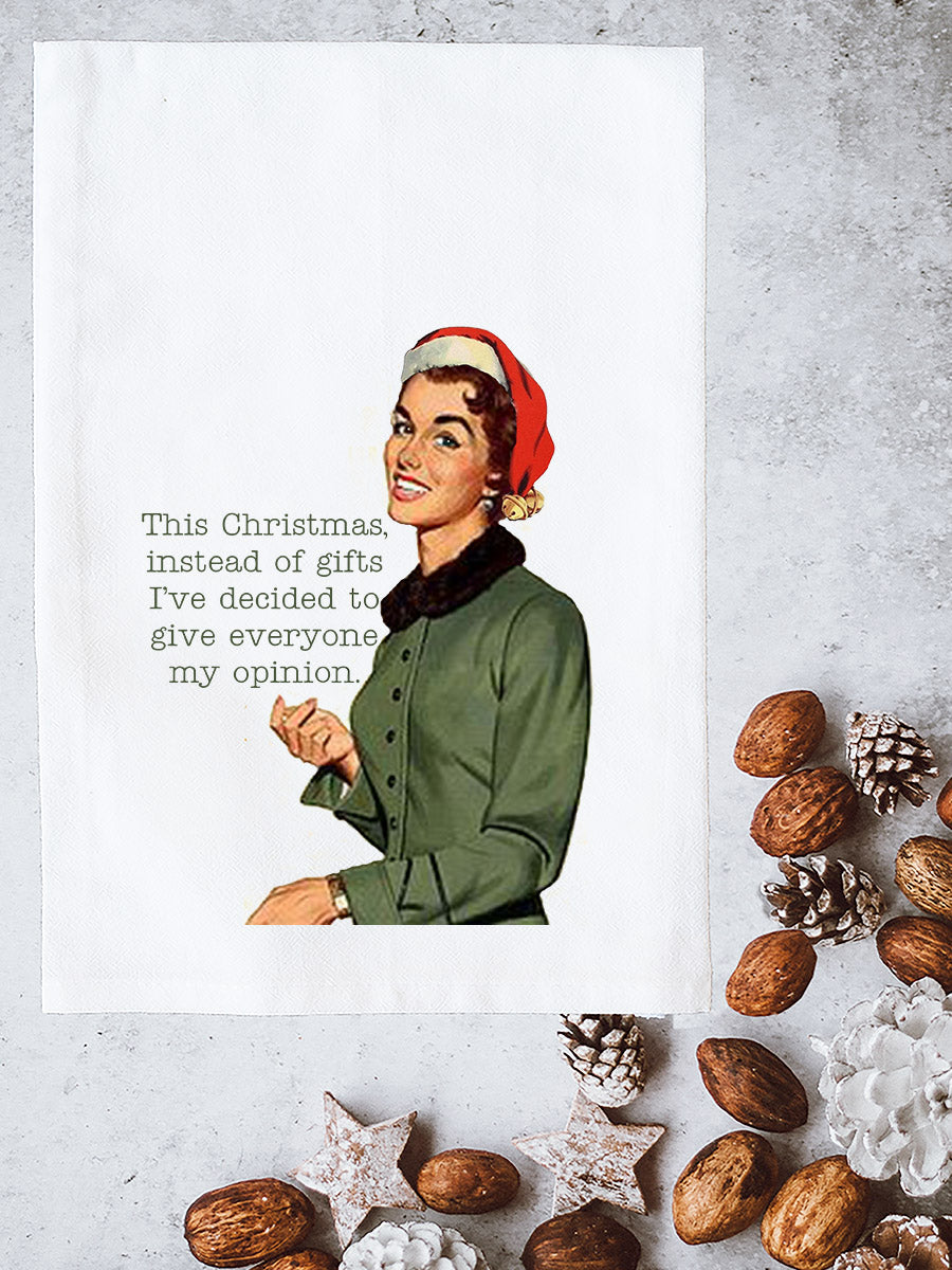 A white kitchen tea towel with a vintage-style illustration of a woman in a Santa hat and green jacket, next to the text 'This Christmas, instead of gifts I've decided to give everyone my opinion.' The towel is styled flat-lay on a gray textured surface with nuts, pinecones, and white star ornaments around it.