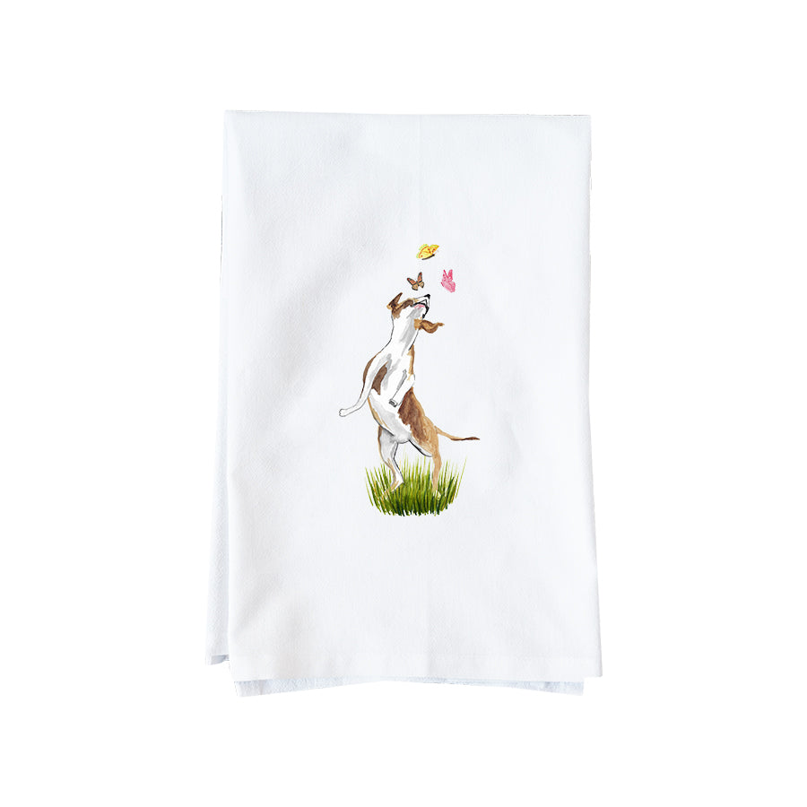 A white kitchen tea towel featuring a watercolor illustration of a white and brown dog standing on its hind legs to catch a butterfly above green grass.