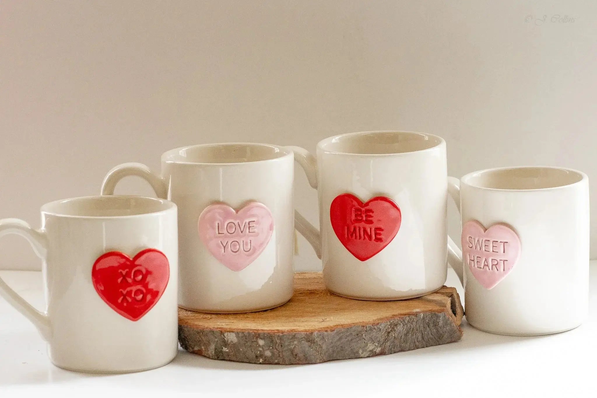 Set of four white ceramic mugs with red and pink heart designs reading 'XOXO,' 'Love You,' 'Be Mine,' and 'Sweet Heart,' displayed on a rustic wood slice for cozy farmhouse decor.