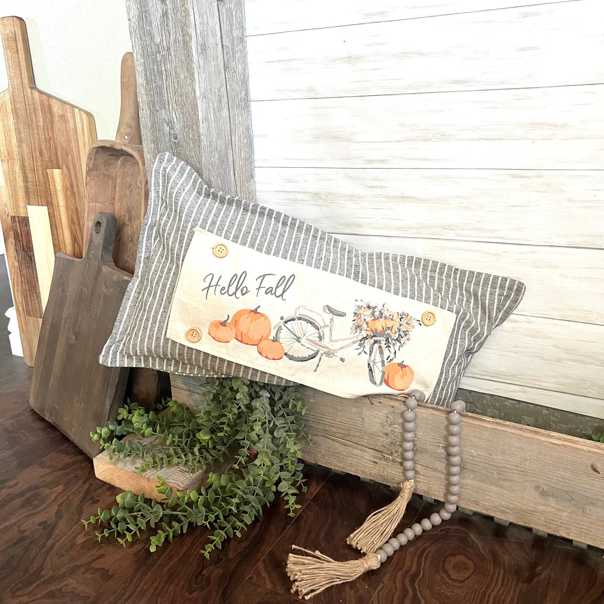 A rectangular striped throw pillow with a 'Hello Fall' autumn illustration, styled on a rustic wooden bench with a wooden bead tassel and green foliage.