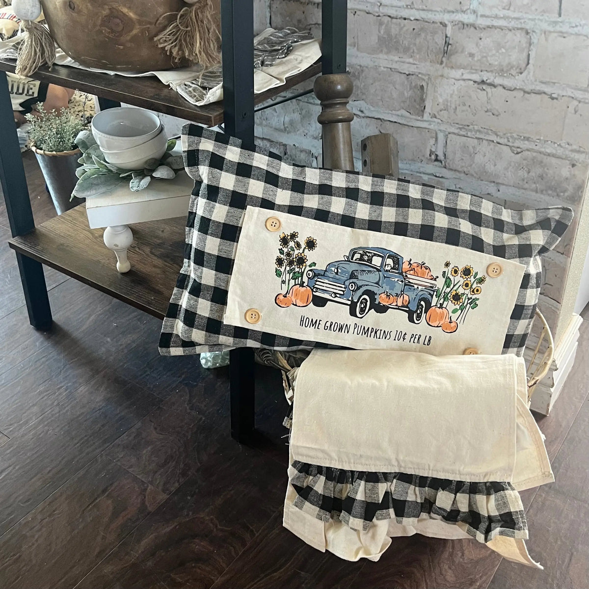 A decorative throw pillow with a black and white gingham border and a central cream panel featuring a vintage blue pickup truck loaded with pumpkins and sunflowers, styled on a wooden bench in a rustic room.