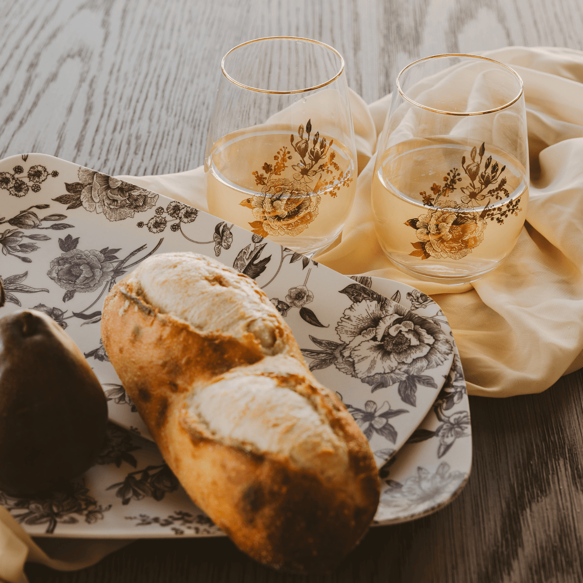 Two bread rolls on a floral plate with two glasses of white wine on a wooden table.