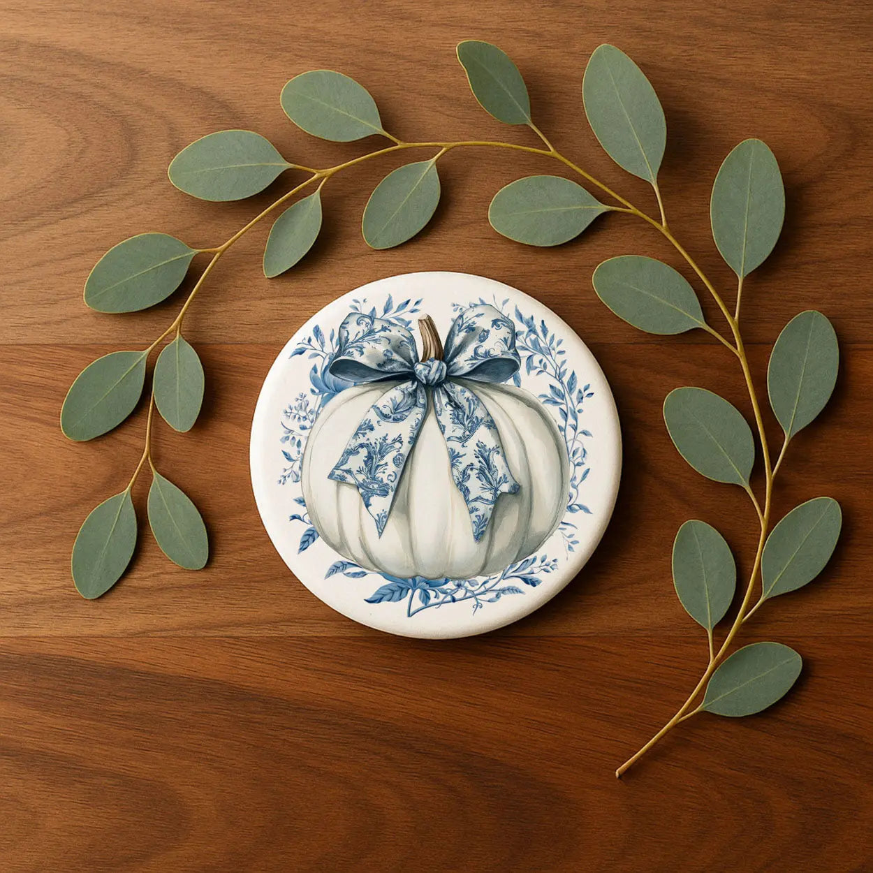 Decorative plate with pumpkin design on a wooden surface with eucalyptus leaves.