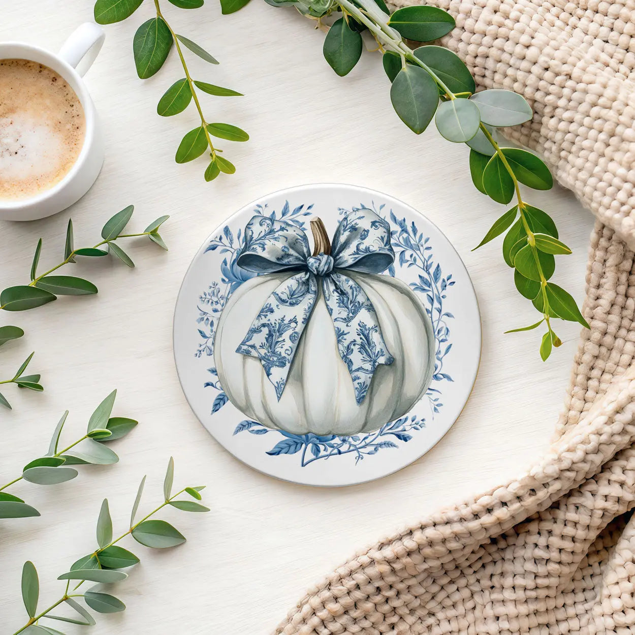 Decorative plate with a pumpkin design and blue bow, surrounded by green leaves and a cup of coffee.