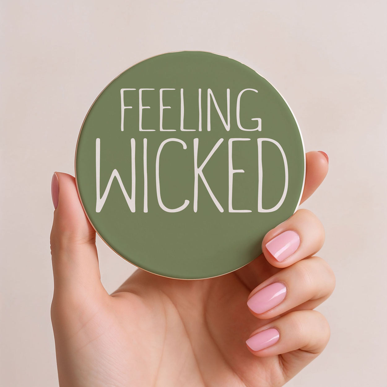 Hand holding a green round object with 'FEELING WICKED' text on a beige background