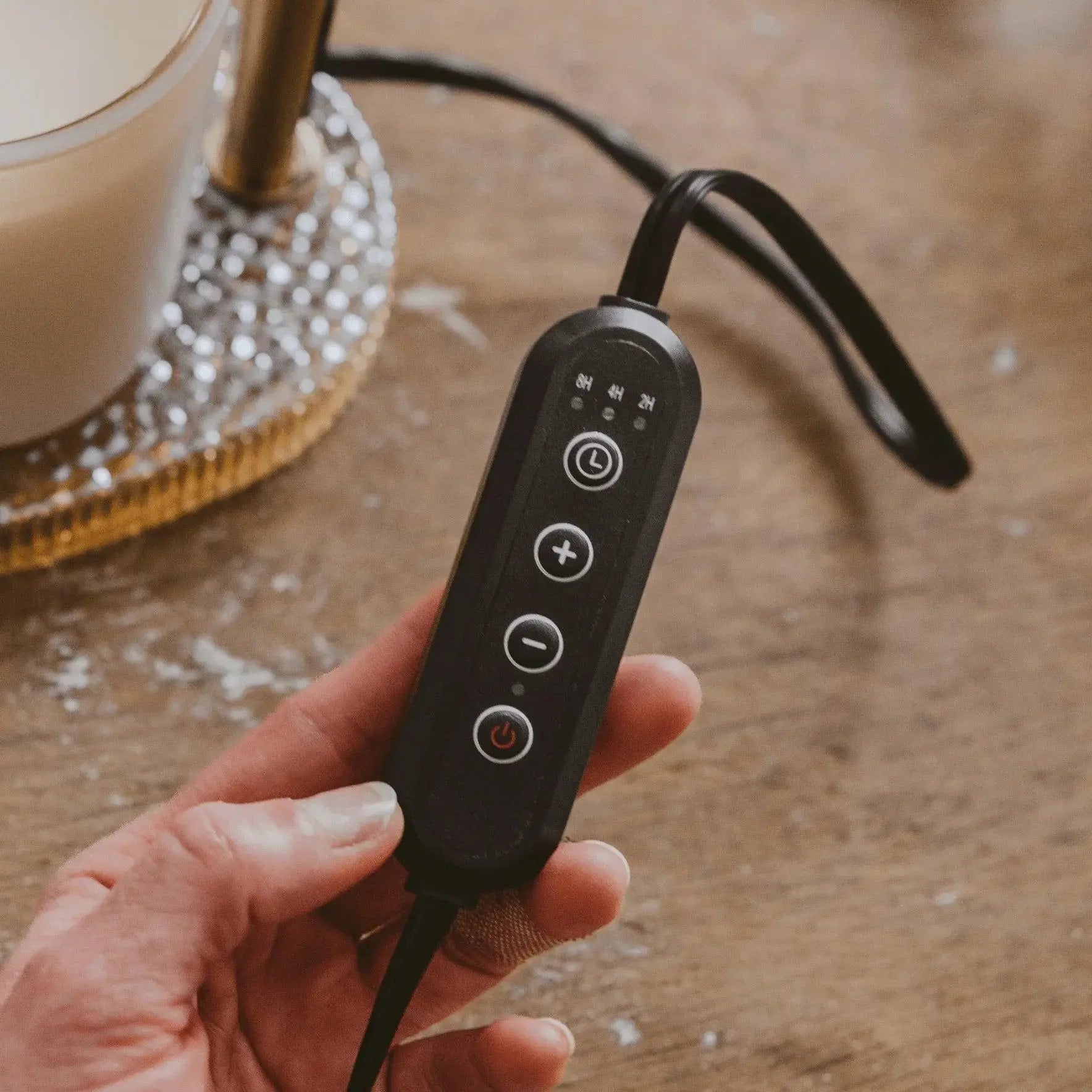 Hand holding black candle warmer control with timer buttons, near cozy farmhouse-style candle on wood table