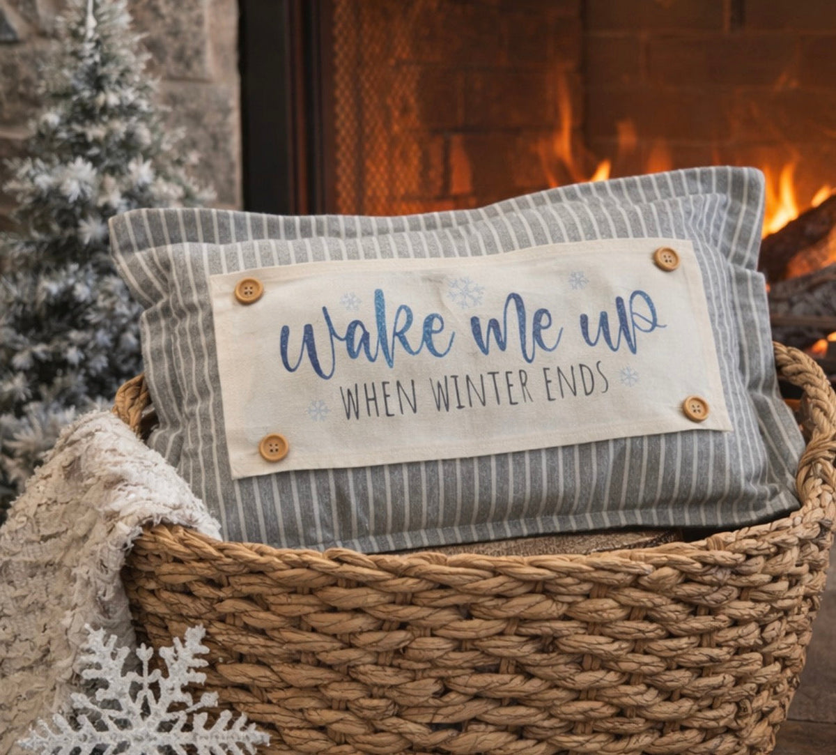 Decorative pillow with 'Wake me up when winter ends' text in a woven basket by a fireplace.