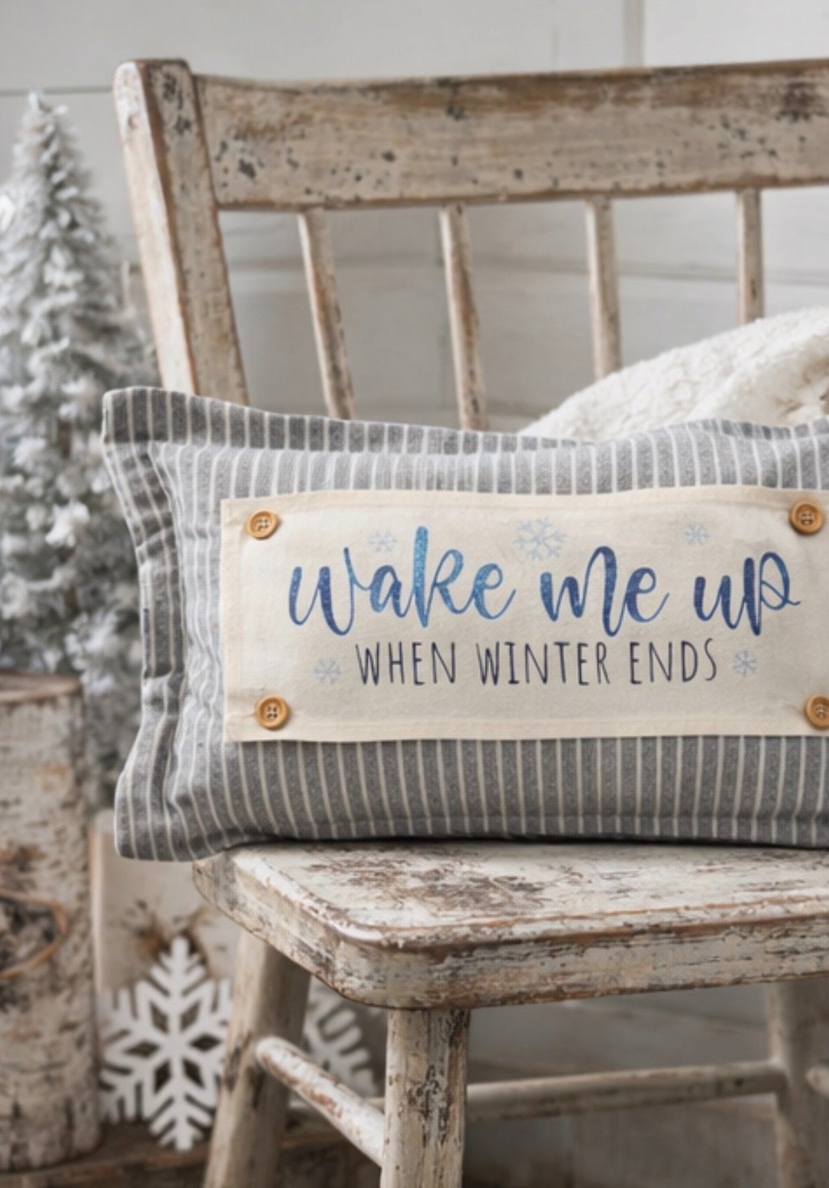 A rectangular decorative throw pillow with a gray and white striped fabric body and a cream center panel reading 'wake me up WHEN WINTER ENDS' in blue lettering, styled on a distressed wooden chair.