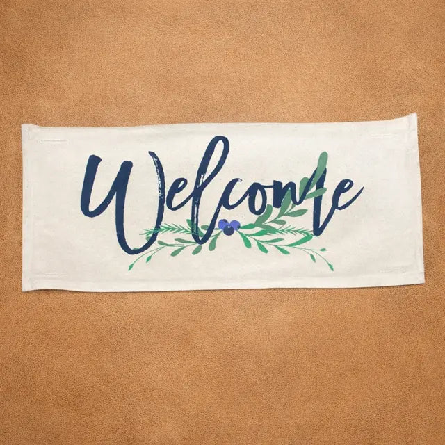 A rectangular cream-colored fabric sign or towel featuring the word 'Welcome' in dark navy blue script, decorated with green leafy branches and a small purple berry, displayed flat against a textured tan background.