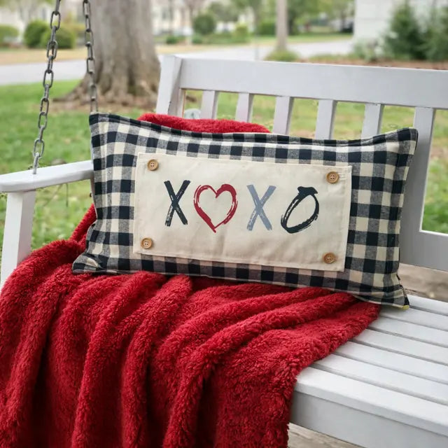Decorative pillow with 'XOXO' text on a red blanket over a white bench outdoors.