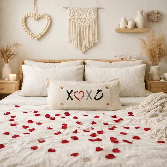 A cozy bedroom scene with a bed dressed in white bedding and scattered red rose petals, featuring a rectangular cream pillow in the center with embroidered 'XOXO' lettering and a heart motif. The room includes macrame wall decor, a wooden headboard, bedside tables, and dried floral arrangements.