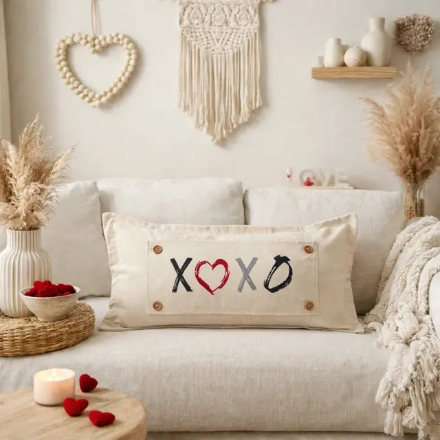 A beige rectangular throw pillow with embroidered 'X', 'O', and 'XO' lettering, styled on a light beige sofa in a cozy, boho-themed living room setting.