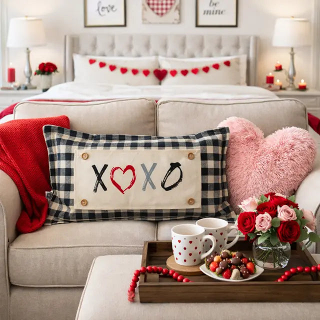 A Valentine's Day themed bedroom and living room scene featuring a beige sofa with a black and white gingham pillow that reads 'XOXO' with a red heart, a pink heart pillow, and a red throw blanket. A coffee table in front holds a wooden tray with red beaded garlands, a white polka-dot mug, cookies, and a small vase of red and pink roses.