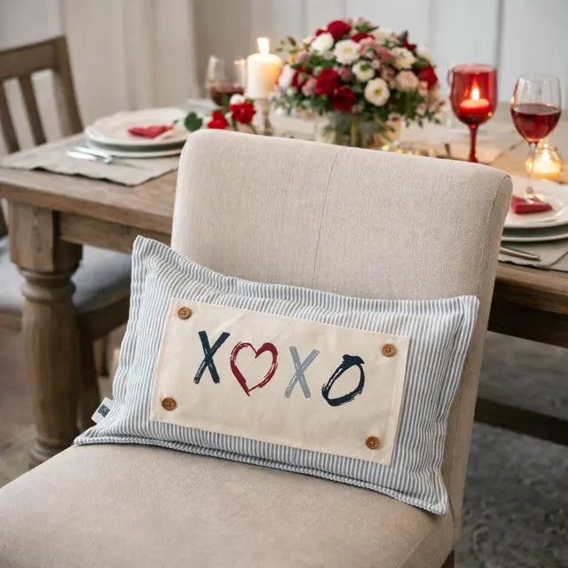A rectangular throw pillow with a striped blue and white border and a cream center panel reading 'XOXO' with a red heart, resting on a beige dining chair in a softly lit dining room.