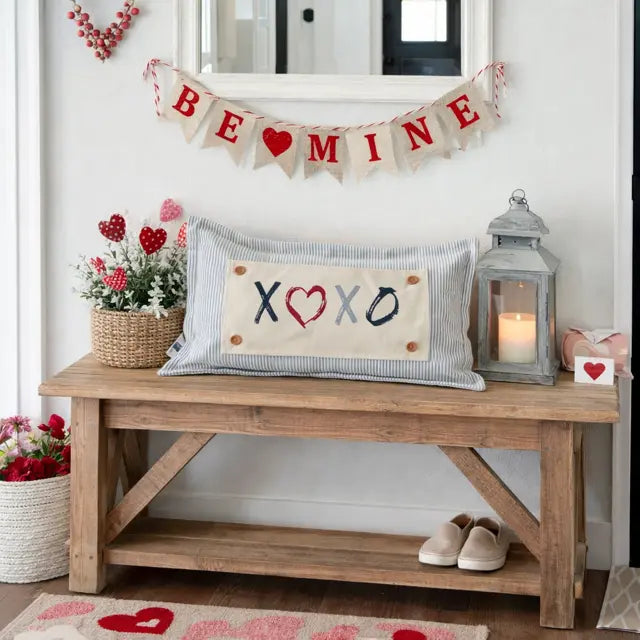 A decorative rectangular throw pillow with a striped blue-and-white border and a cream center panel reading “XOXO” with a red heart, styled on a wooden bench in a Valentine’s-themed room. A “BE MINE” banner hangs above, with flowers, a lantern, and shoes nearby.