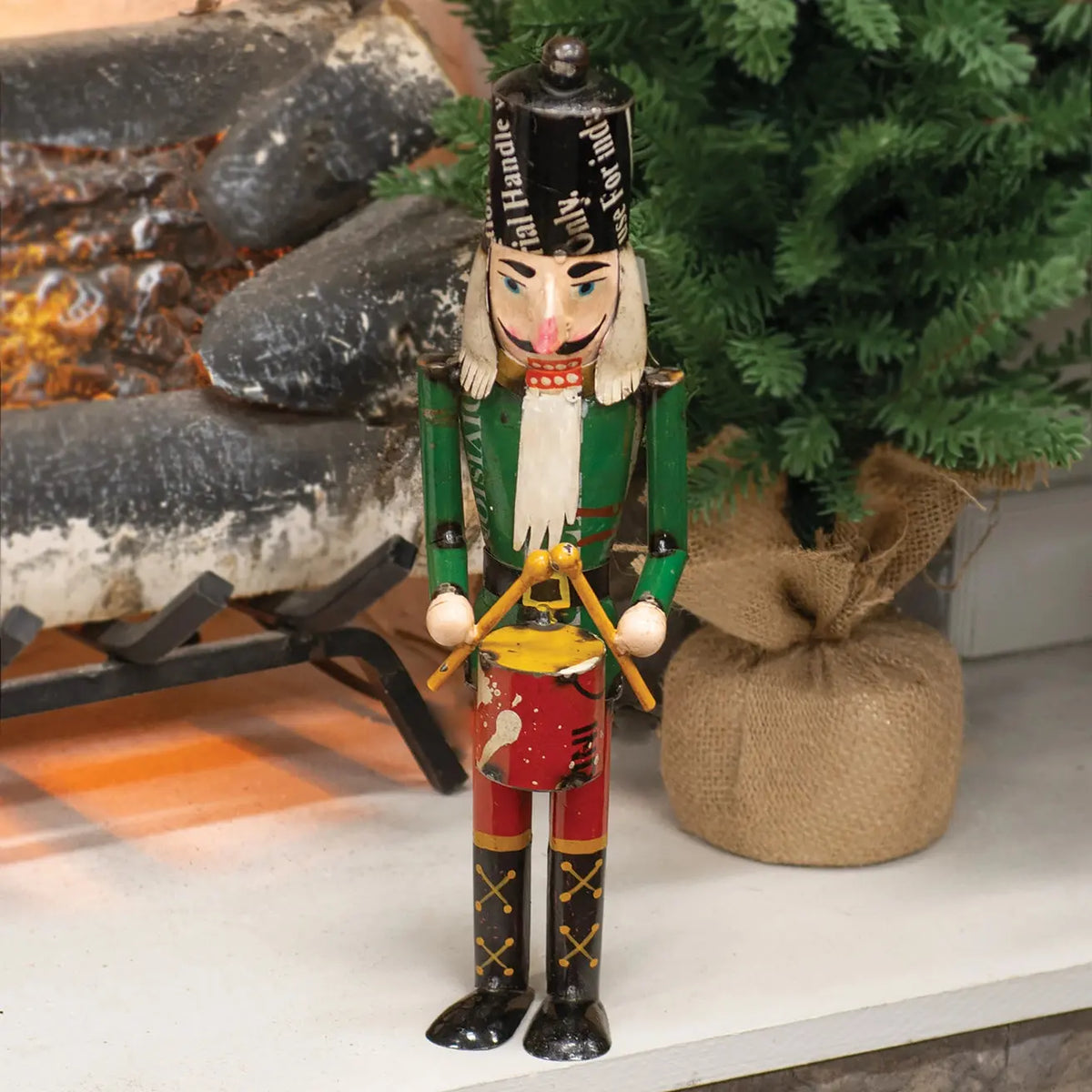 A painted nutcracker figurine stands on a white surface in a holiday setting, with a burlap sack and evergreen branches behind it.