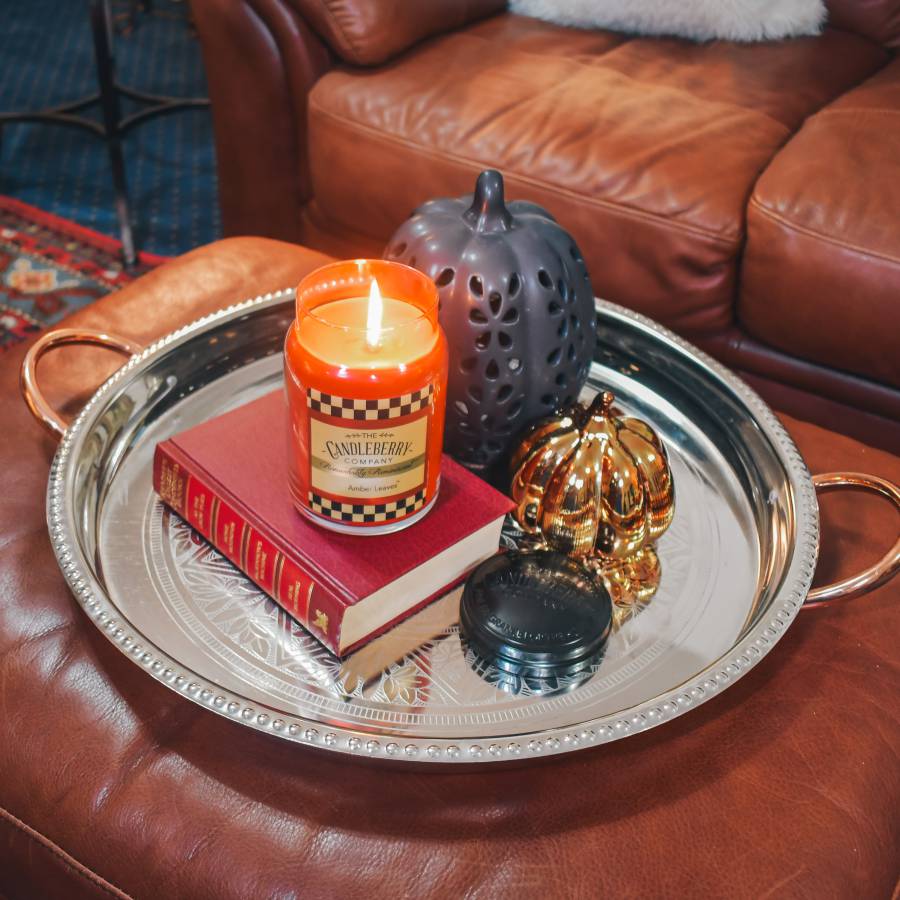 A lit orange candle in a jar with a checkered label reading Candlelight, styled on a silver tray with a red book, a dark perforated pumpkin, a gold pumpkin, and a black lid on a brown leather ottoman.