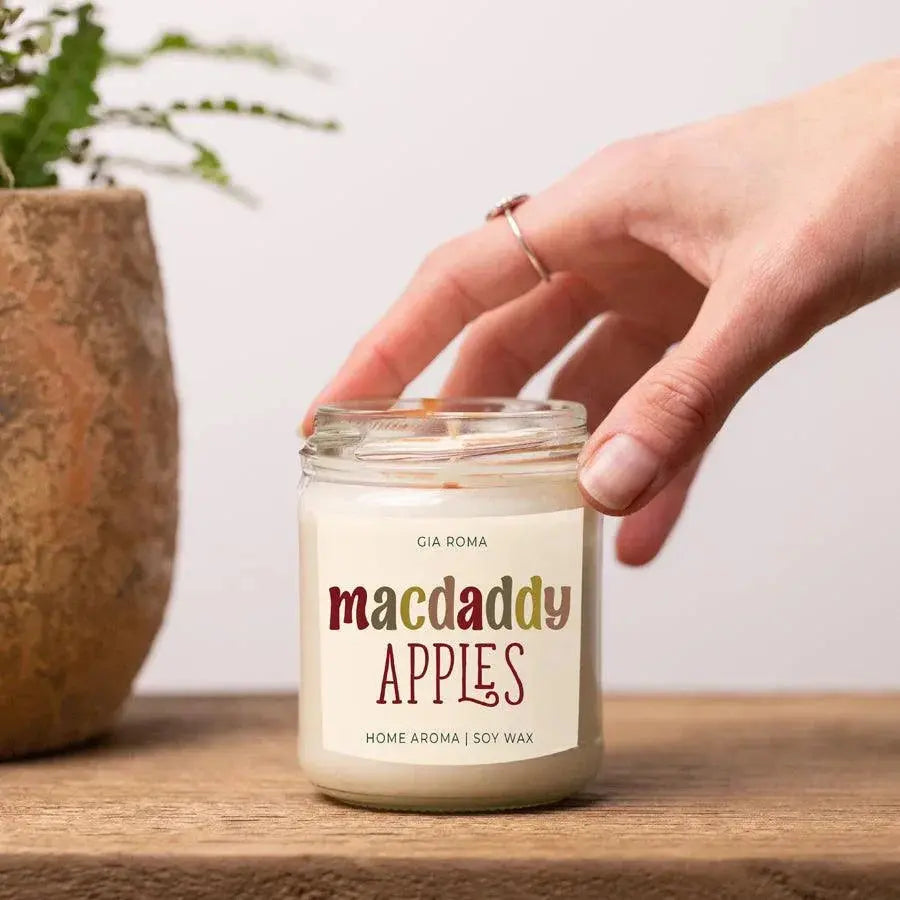 Candle labeled 'macdaddy APPLES' held by a hand on a wooden surface with a plant in the background.