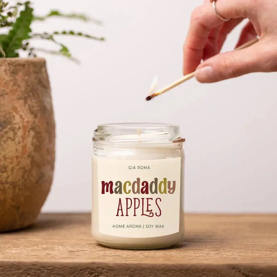 Candle labeled 'macdaddy APPLES' being lit with a match on a wooden surface.