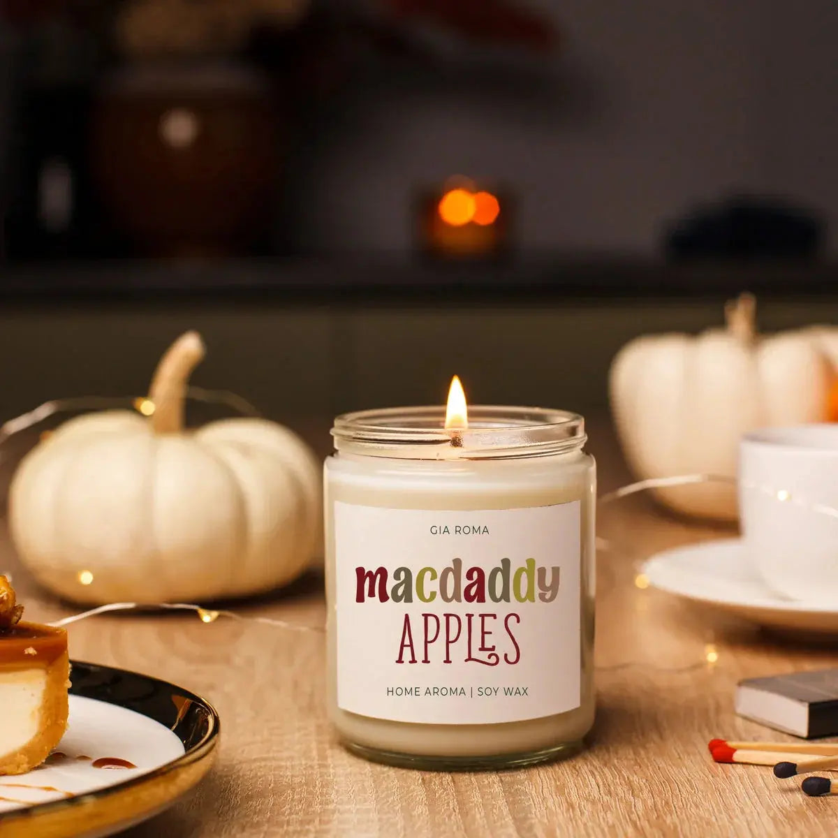 Candle labeled 'macdaddy APPLES' on a table with pumpkins and a cup in the background