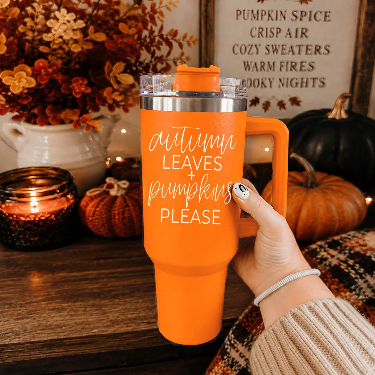 Orange mug with text held by a hand, surrounded by autumn decorations.
