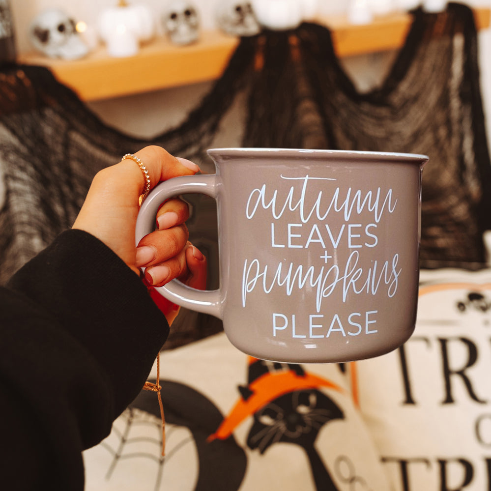 Hand holding a mug with 'Autumn leaves + pumpkins please' text against a Halloween-themed background.