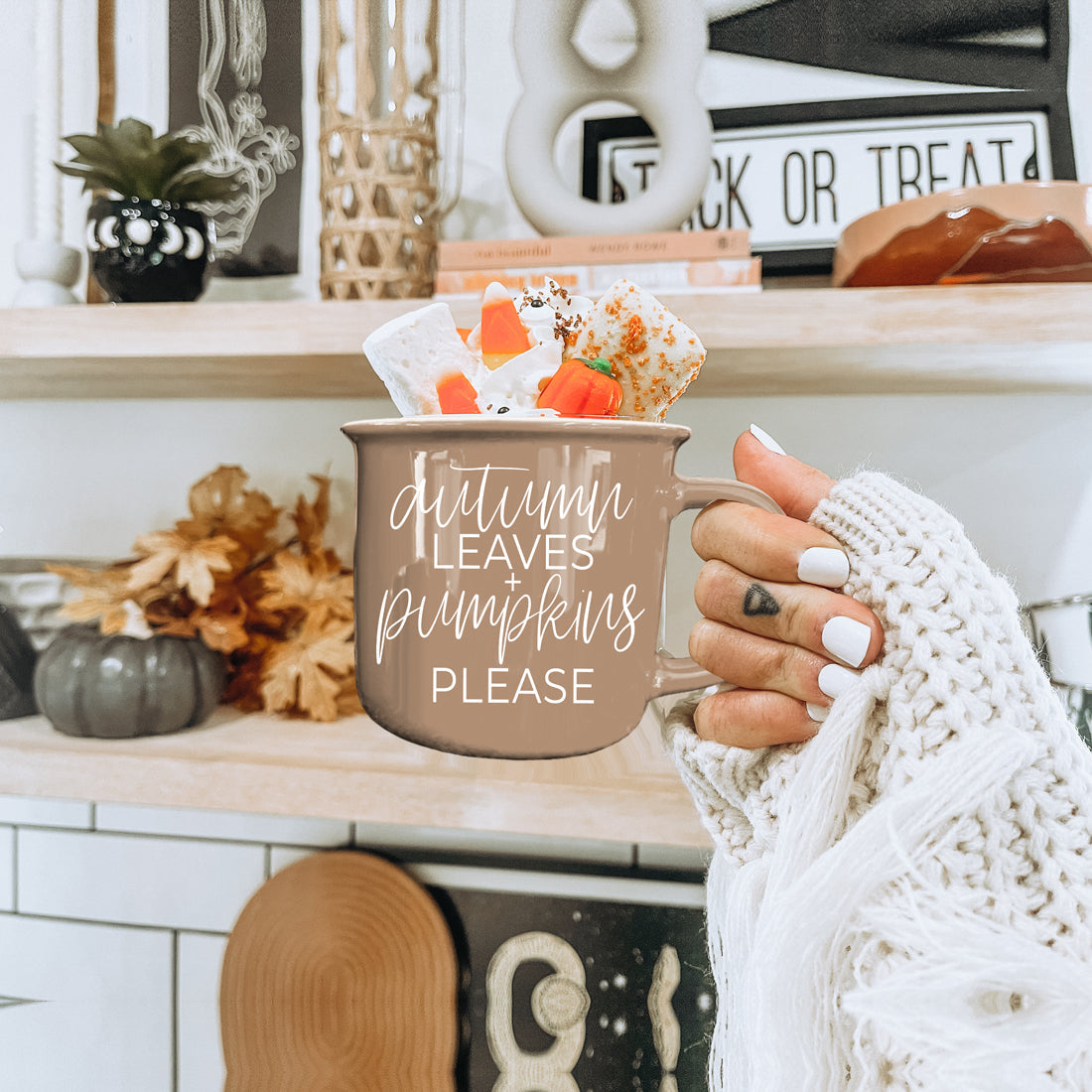 Person holding a mug with 'Autumn leaves pumpkins please' text in a cozy kitchen setting.